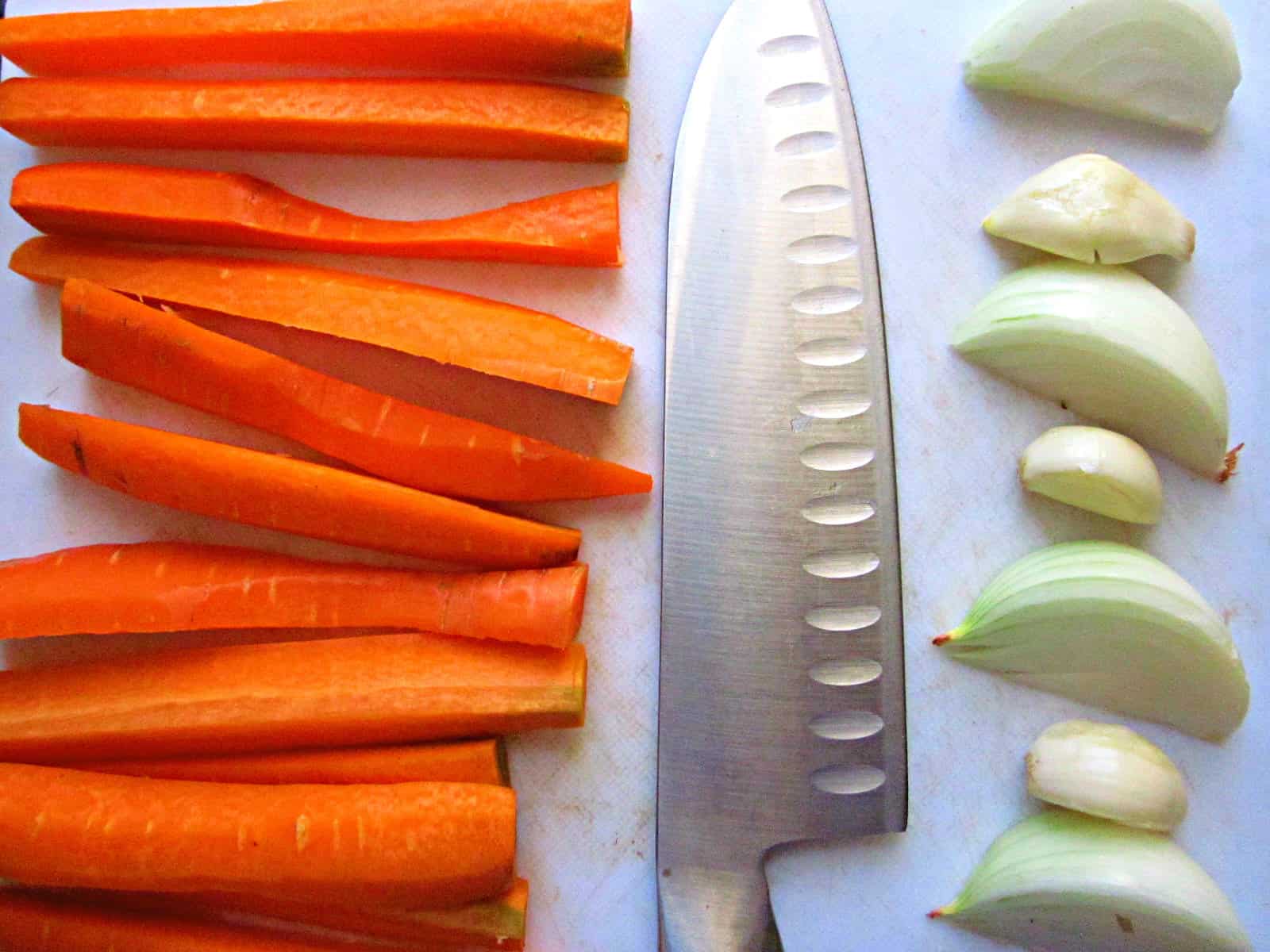 Pickles' ingredients Carrot sticks, quartered onion pieces, garlic cloves, and a large chef's knife are arranged neatly on a white cutting board, ready to be prepared into delicious lacto-fermented vegetables.