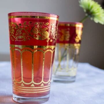 A glass of tea in an ornate red and gold glass, with another glass holding a flower and a hint of strawberry compote in the background.