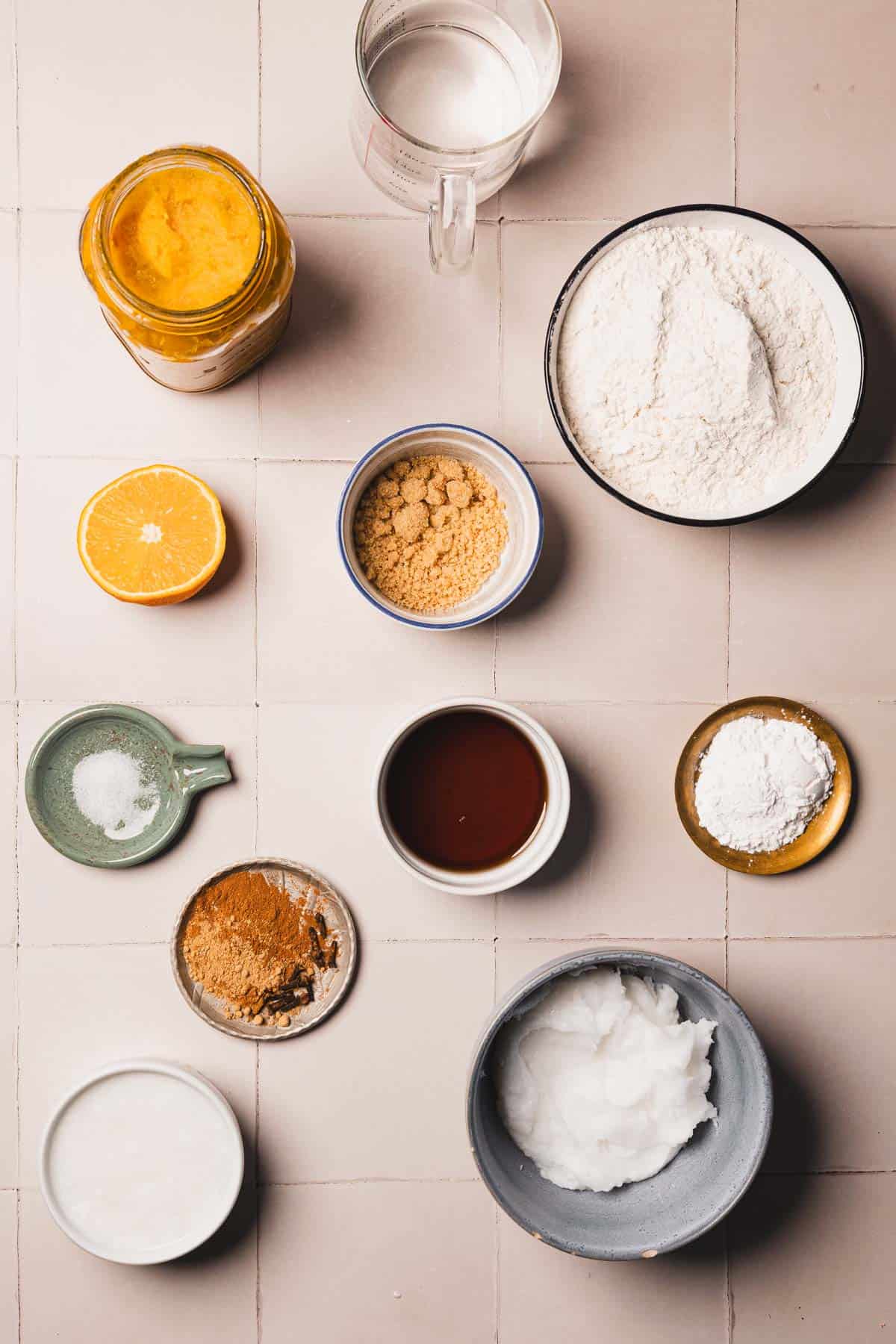 Various baking ingredients in bowls and jars, including flour, coconut oil, spices, lemon, and syrup on a tiled surface-perfect for creating a comforting hubbard squash pie.