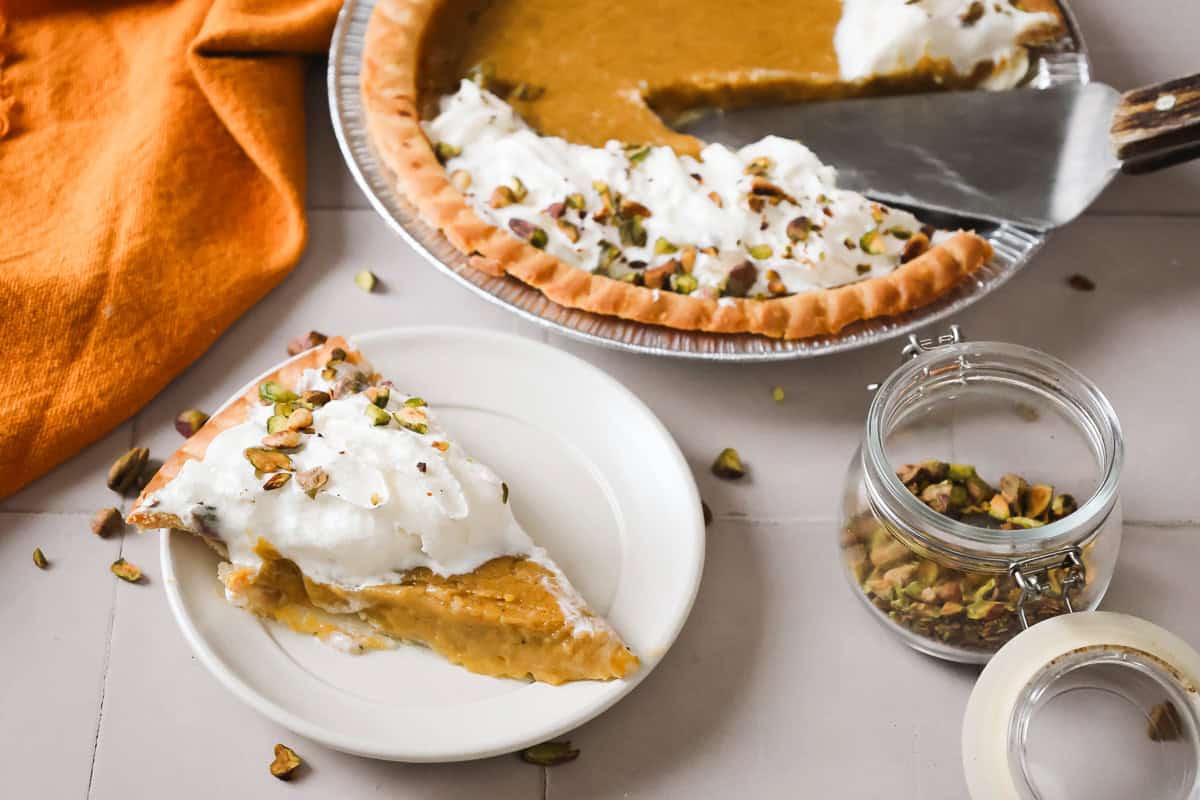A slice of hubbard squash pie with whipped cream and chopped pistachios on a plate, next to the remaining pie and a jar of pistachios.