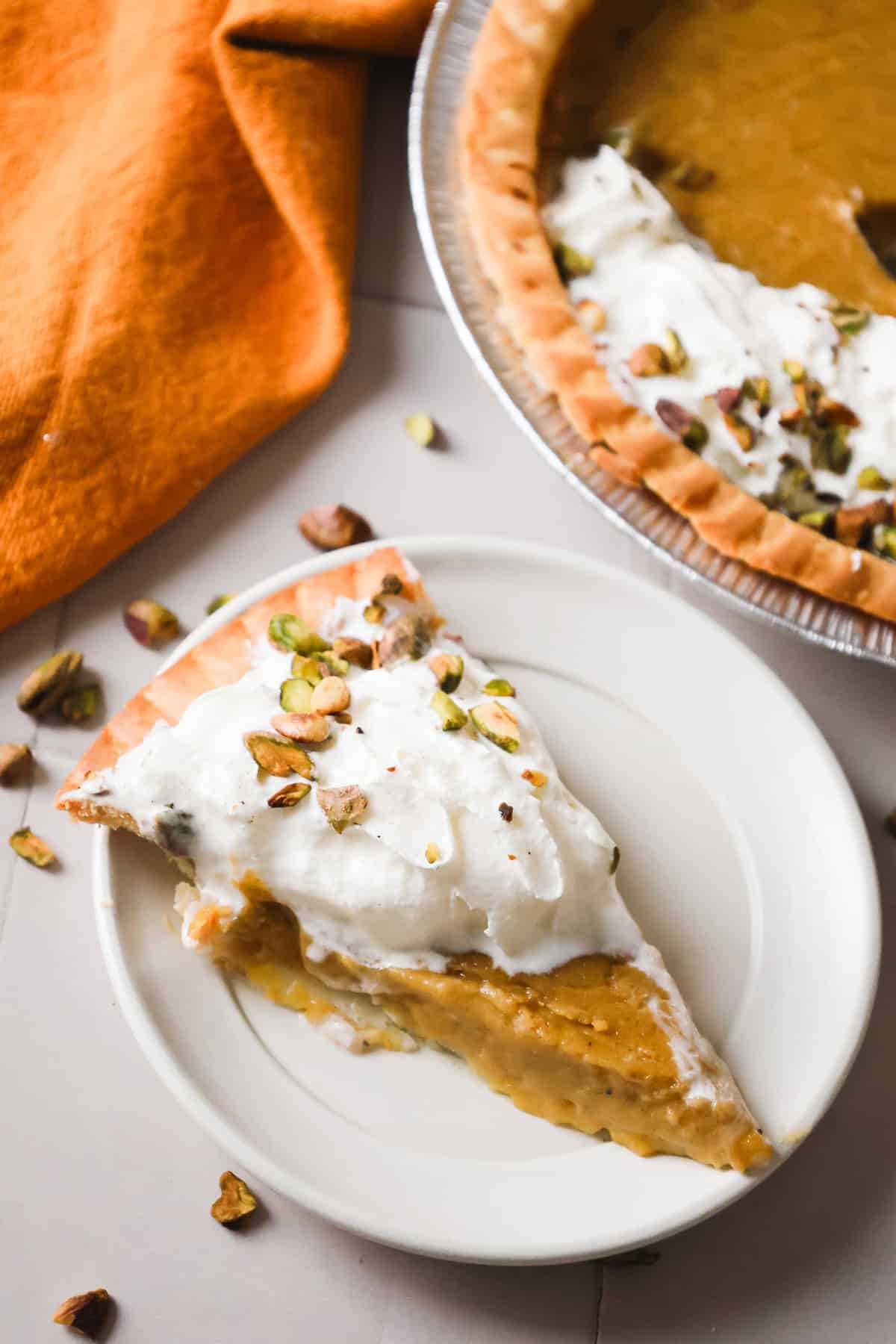 A slice of hubbard squash pie with whipped cream and chopped pistachios on a white plate, next to the rest of the pie.