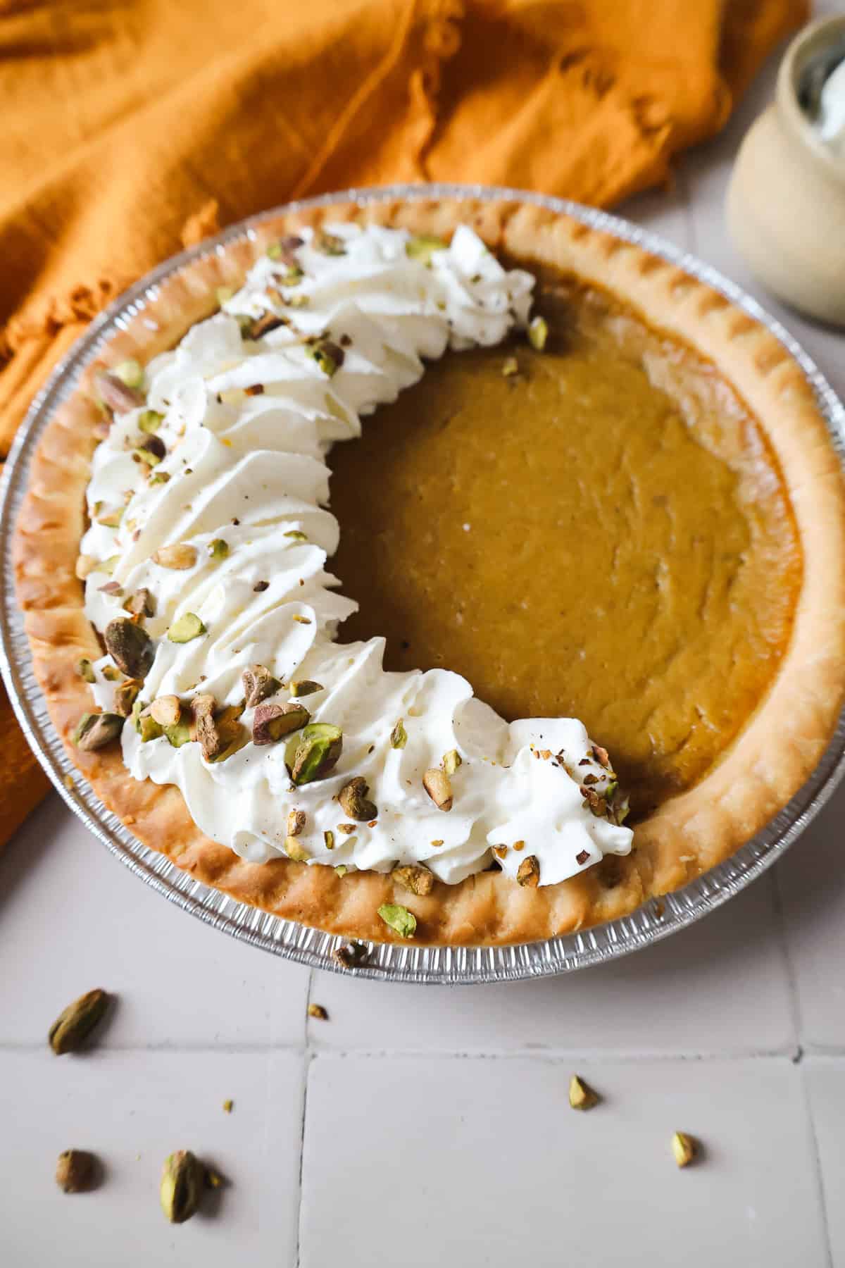 A hubbard squash pie topped with a crescent of whipped cream and chopped pistachios rests on a tiled surface.