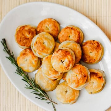 A white plate with a plate of mini puff pastry pockets filled with savory pies and garnished with rosemary sprigs.