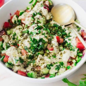 A bowl of fresh Israeli chopped salad topped with creamy dressing and sprinkled with herbs. The salad includes tomatoes, cucumbers, and red onions, and is accompanied by a pair of gold utensils. Parsley leaves and tomatoes are visible in the background.