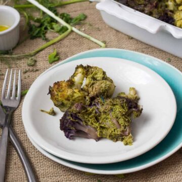 A white plate with roasted mixed-colored cauliflower in green herb sauce rests on a blue-bordered dish. Nearby are a fork and knife, a small dish holding vibrant yellow sauce, scattered herbs, and a white baking dish filled with more roasted cauliflower on a burlap surface.