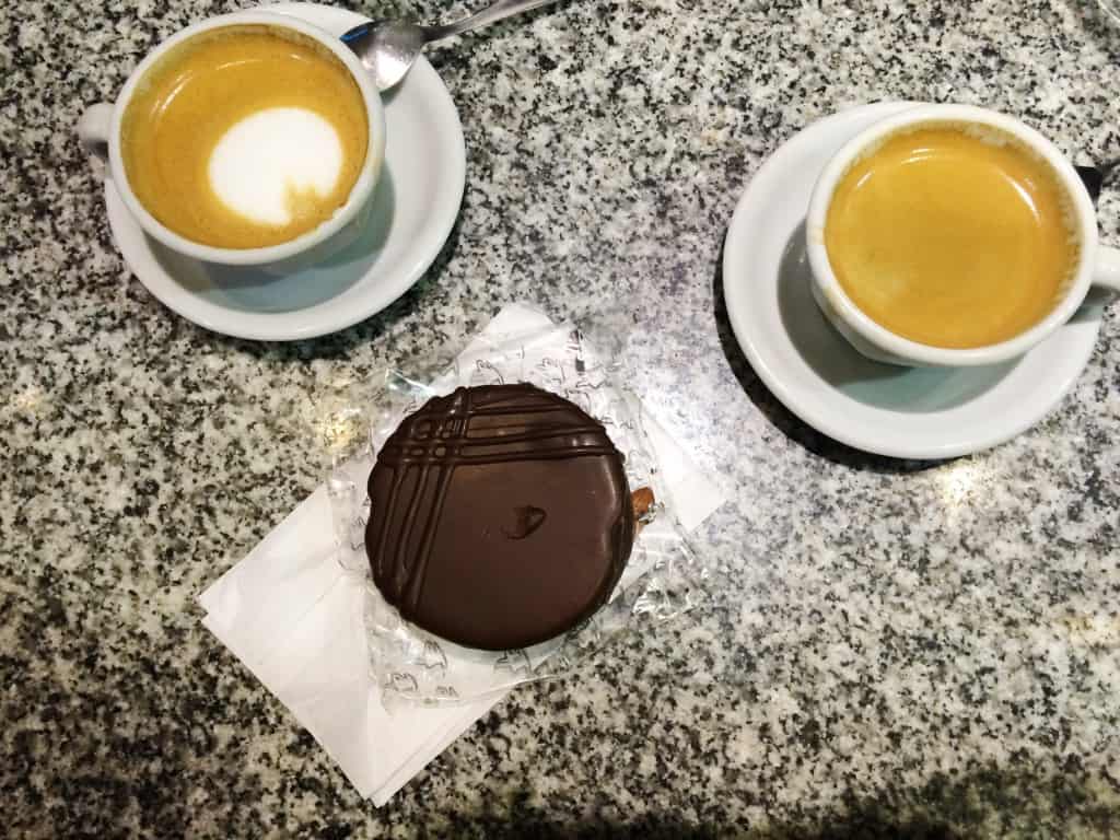 Two cups of coffee, one topped with milk foam and the other black, sit on a granite countertop. Nestled between them is a chocolate-covered cookie on a paper wrapper—gluten-free alfajores to complement your perfect coffee break.