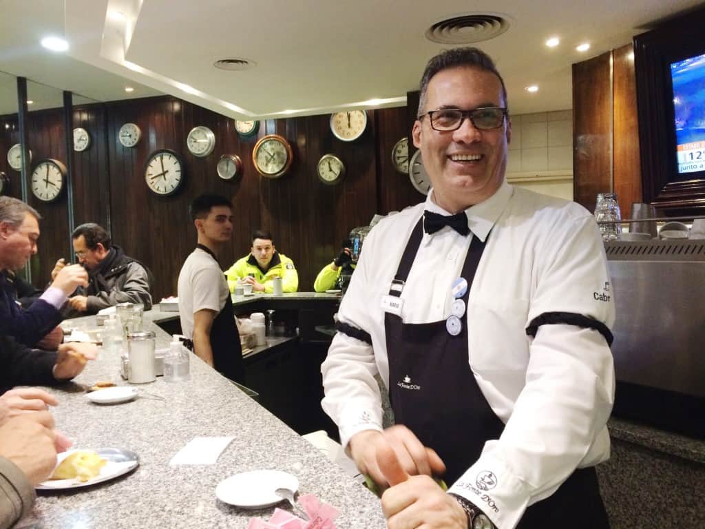 A smiling bartender in glasses and a bow tie stands behind a cafe counter, offering gluten-free alfajores to the patrons seated there. Multiple wall clocks show different times behind him, and a television screen flickers to the right.