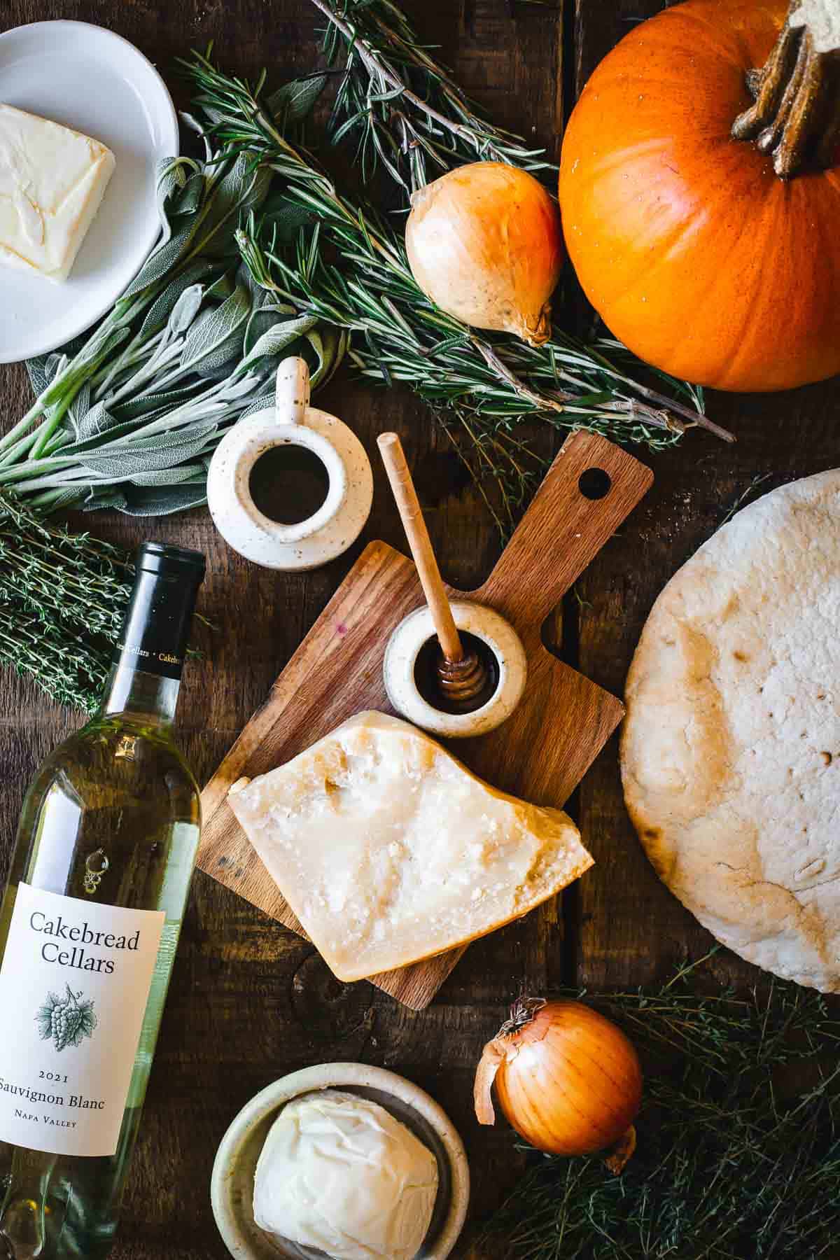 A bottle of wine, cheese, and herbs on a wooden table. Ingredients for pumpkin pizza.