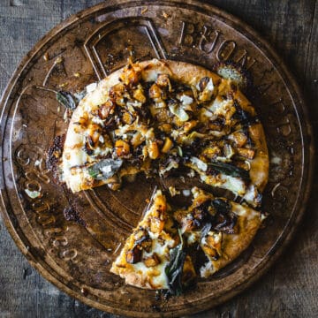 A pizza on a wooden plate with a slice taken out, topped with garlic, onions and sage.