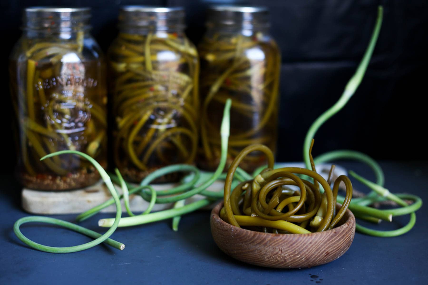 Two jars of garlic scape pickles and a wooden bowl.
