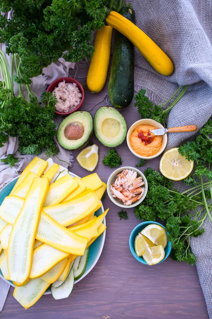 A variety of ingredients are laid out on a table: sliced yellow squash, whole zucchini ready for transformation into crab-stuffed zucchini, avocados, lemon halves, parsley, and small bowls filled with hummus, shredded fish, and seasoning. A gray cloth is partially visible in the background.