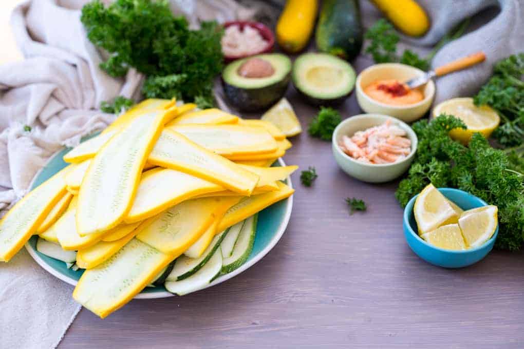 A plate with sliced yellow and green zucchini is surrounded by fresh parsley, avocado, lemon wedges, and small bowls of various ingredients on a wooden surface. In the background, a zucchini hints at delicious possibilities like crab-stuffed zucchini or vibrant zucchini rolls.