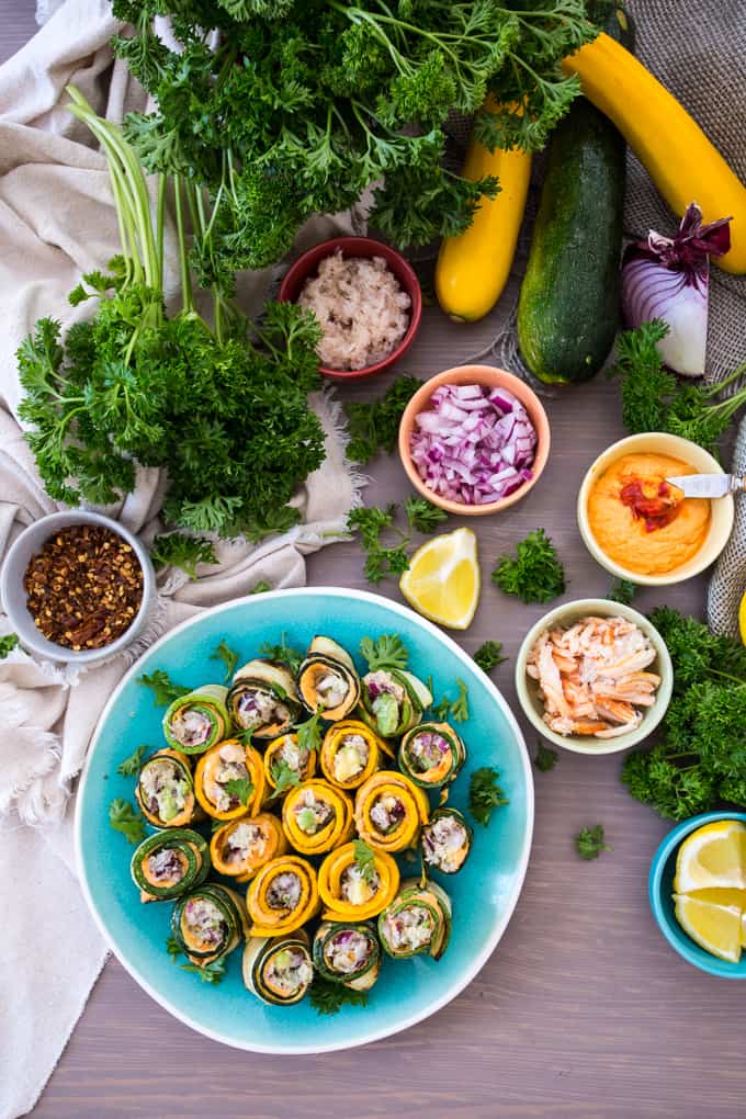 A plate of zucchini rolls filled with various ingredients is surrounded by fresh parsley, sliced onion, squash, a lemon wedge, and small bowls of dipping sauces and seasonings on a wooden surface.