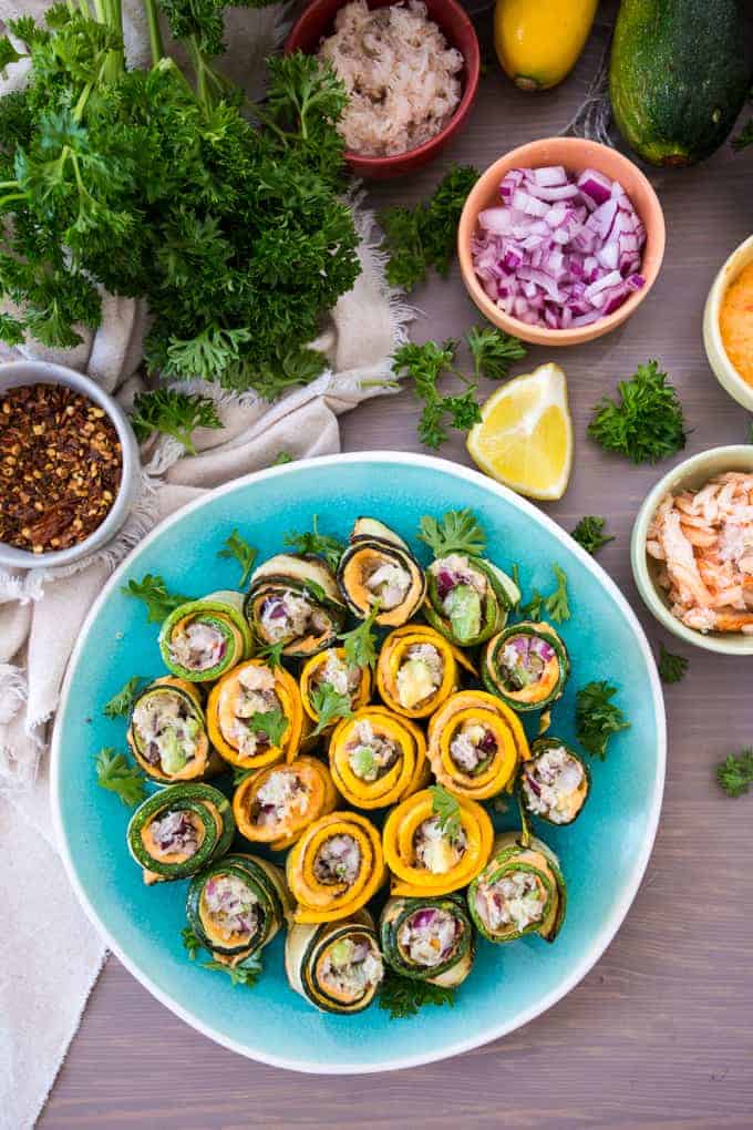 A plate of zucchini and yellow squash rolls with crab-stuffed fillings, garnished with parsley, sits on a table. Surrounding the plate are small bowls containing red onion, shredded ingredients, red pepper flakes, a lemon wedge, and fresh parsley.