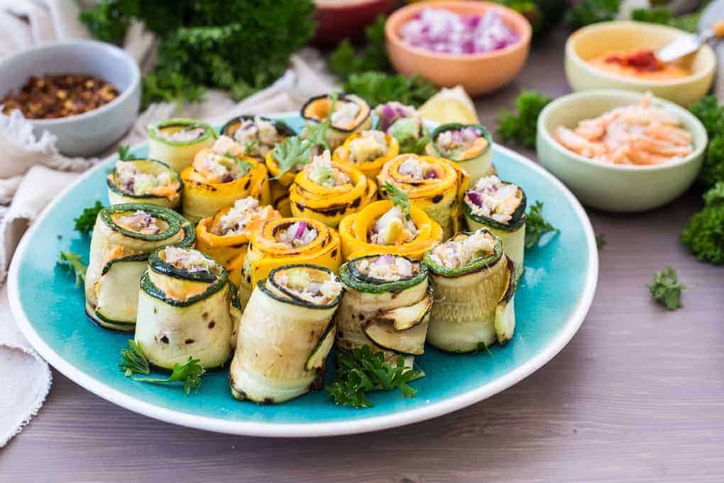 A plate of zucchini and yellow squash rolls filled with a creamy mixture, garnished with parsley. The plate sits on a wooden surface, surrounded by small bowls containing red onion, shrimp, spices, and hints of crab stuffed zucchini to elevate its delectable charm.