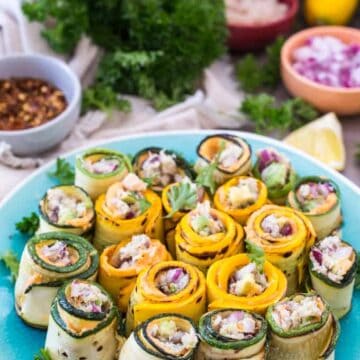 A plate of zucchini rolls, delicately filled with crab, rests on a table. Garnished with parsley, the colorful dish is surrounded by bowls of spices and a lemon wedge, with fresh greens in the background, offering a fresh twist to your favorite snow crab recipe.