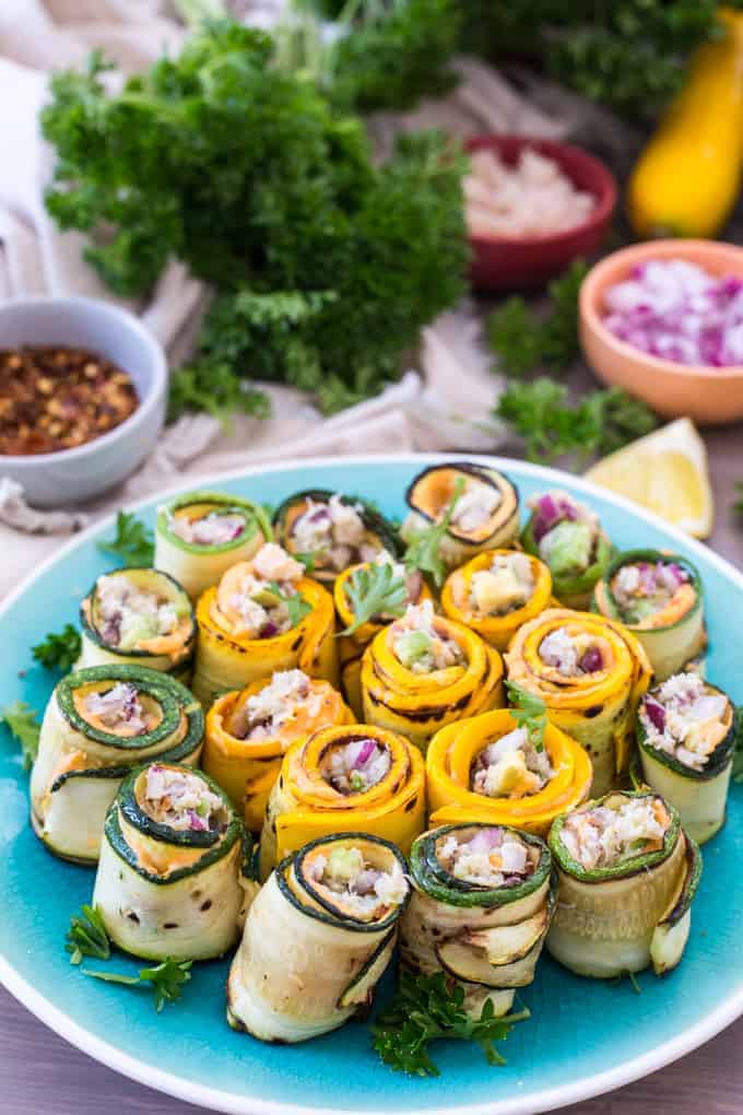 A plate of zucchini rolls filled with a vibrant mixture of ingredients, reminiscent of a crab stuffed zucchini delight. The rolls are arranged on a turquoise plate, garnished with sprigs of herbs. Nearby, small bowls hold spices, diced onions, and leafy greens.