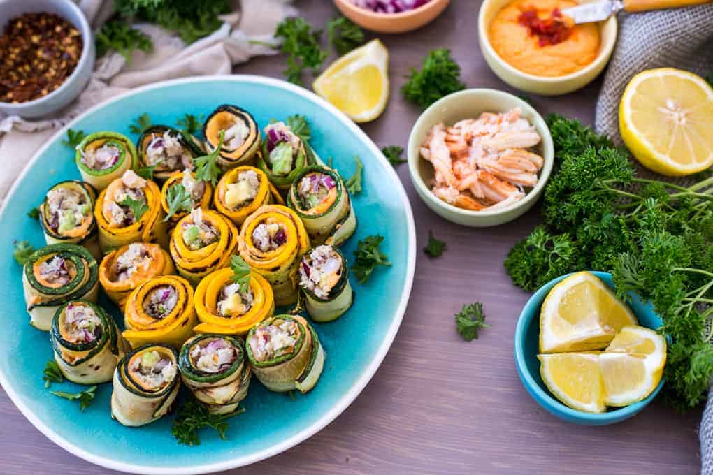 A plate of colorful zucchini rolls garnished with herbs sits on a table. Surrounding the plate are lemon wedges, fresh parsley, and bowls containing various ingredients such as a spread, shredded meat, and chopped vegetables. Perfect for those seeking a twist on the classic snow crab recipe.