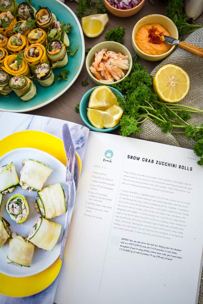 A cookbook open to a snow crab recipe for Zucchini Rolls sits on a table. Nearby are prepared rolls, lemon halves, herbs, and small bowls with ingredients like crab and sauce. A fork is placed on the open book.