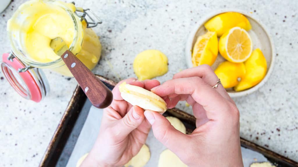 Hands holding a cookie with lemon filling above a baking tray of gluten-free alfajores. A knife rests on a jar of lemon curd, while a bowl with sliced lemons sits nearby on the speckled countertop.