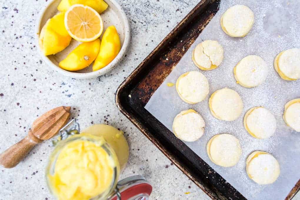 A baking tray lined with parchment paper holds unbaked lemon cookies, reminiscent of gluten-free alfajores. Nearby, a glass jar contains lemon curd. A small bowl with sliced mango and a lemon wedge sits beside a wooden juicer on the speckled countertop.