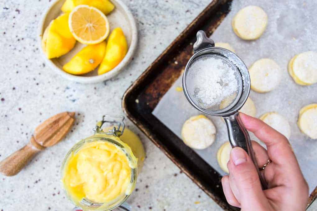 A hand delicately holds a sifter, dusting gluten-free alfajores with powdered sugar over unbaked cookies on a sheet. Nearby, a jar of lemon curd, lemon wedges and zest, and a wooden reamer sit on the speckled countertop.