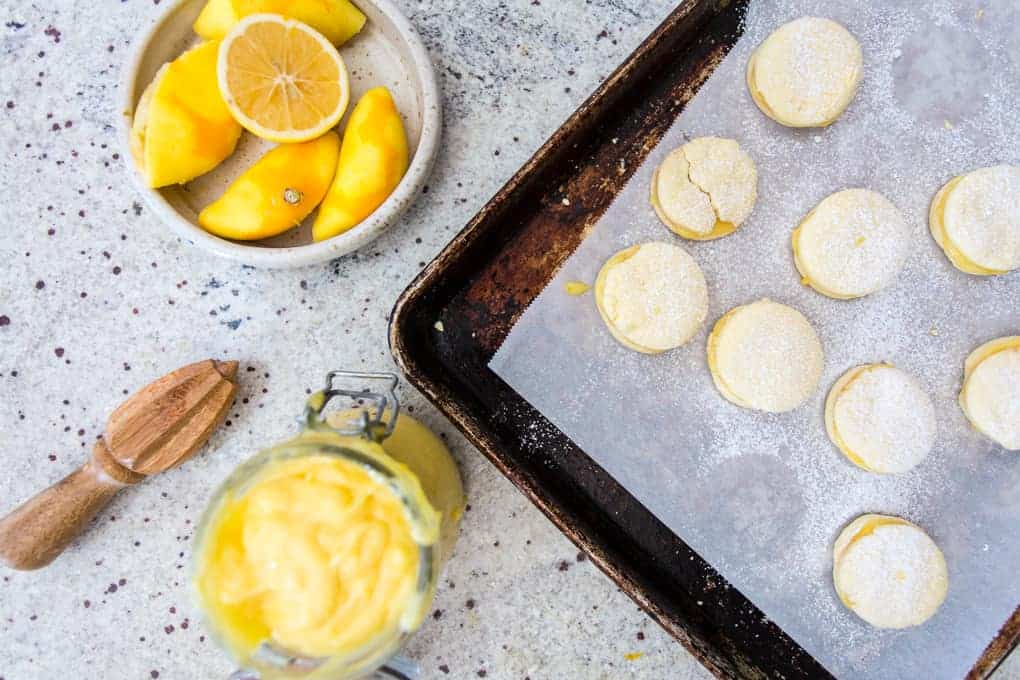 A baking tray with several lemon cookies dusted with powdered sugar sits on a speckled countertop. Nearby, a jar of lemon custard, a citrus reamer, and a plate with lemon wedges accompany gluten-free alfajores, adding a delightful twist to the scene.