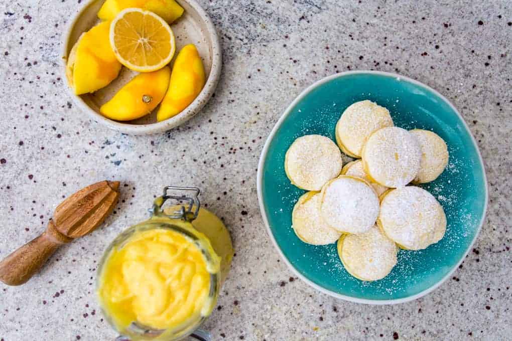 A top-down view of lemon gluten-free alfajores dusted with powdered sugar in a turquoise bowl. Nearby, a jar of lemon curd, sliced lemons and peaches on a plate, and a wooden citrus juicer are arranged on the speckled countertop.