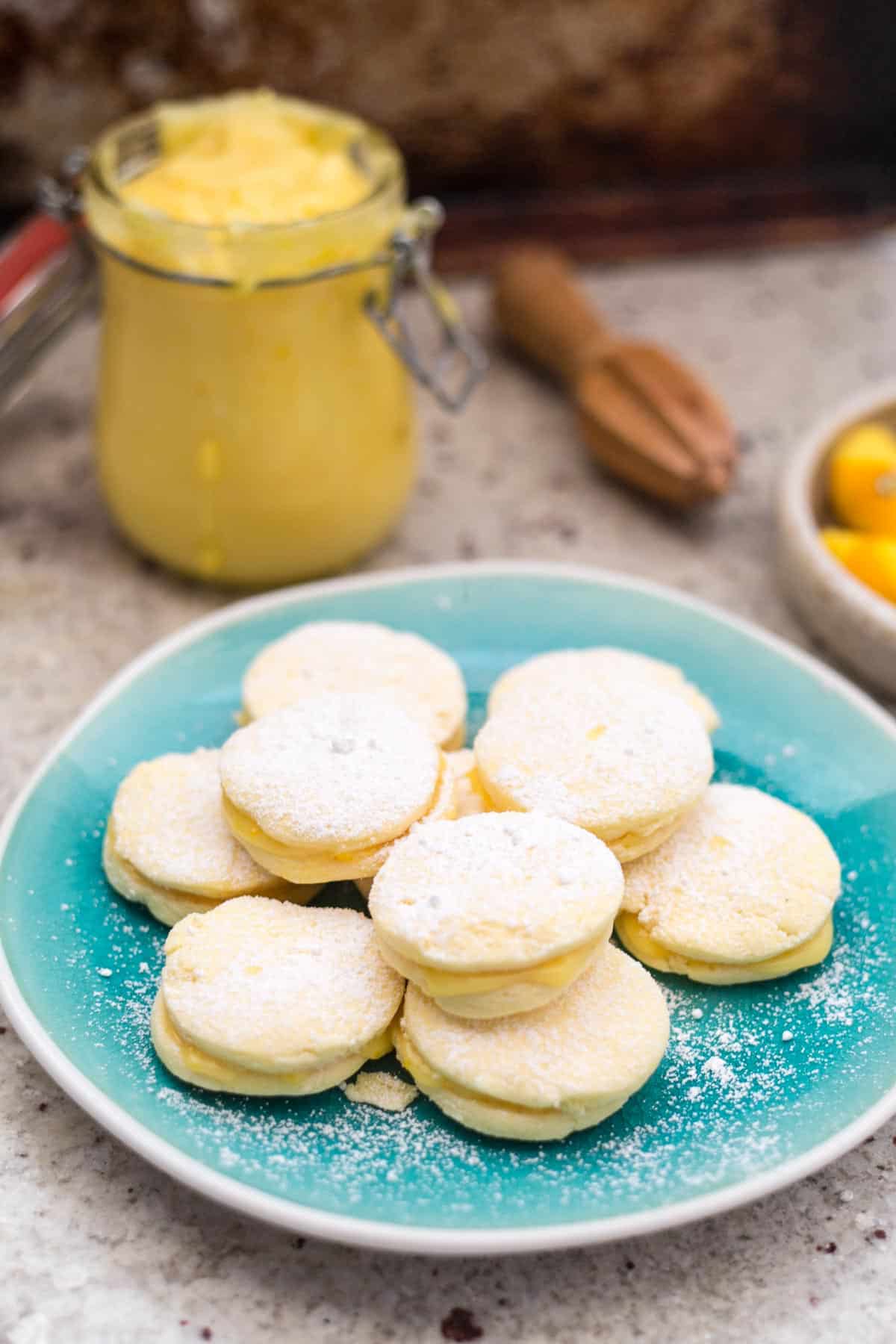 A teal plate holds a stack of round, powdered sugar-dusted gluten-free alfajores. Behind is a jar of yellow cream with a lid, a wooden utensil, and a glimpse of fruit in a bowl, all set on a textured countertop.