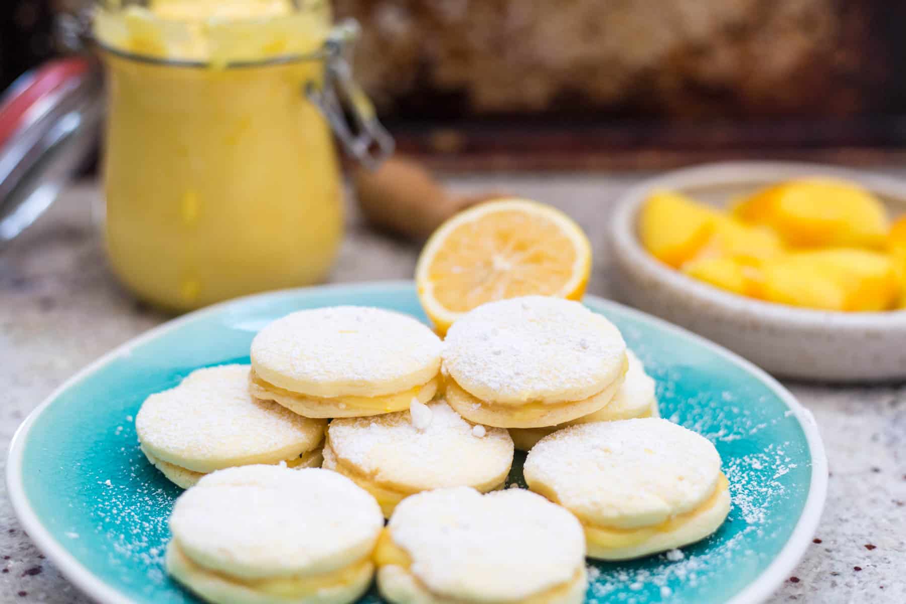 A teal plate displays several gluten-free alfajores, their lemon cream centers dusted with powdered sugar. Behind the plate, a jar of lemon curd, half a lemon, and a bowl of sliced peaches complete the scene against a softly blurred background.