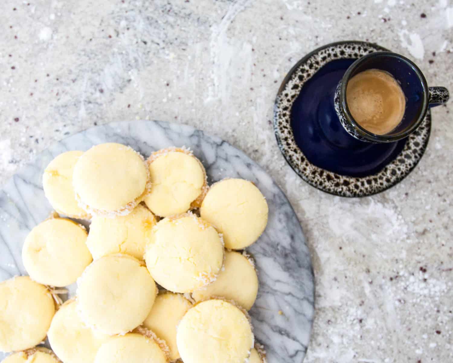 A pile of small, round, yellow pastries that may be gluten-free alfajores sits on a marble slab. The sugar-coated delights are paired with a black cup on a saucer containing what seems to be espresso, placed on a speckled, light-colored surface.