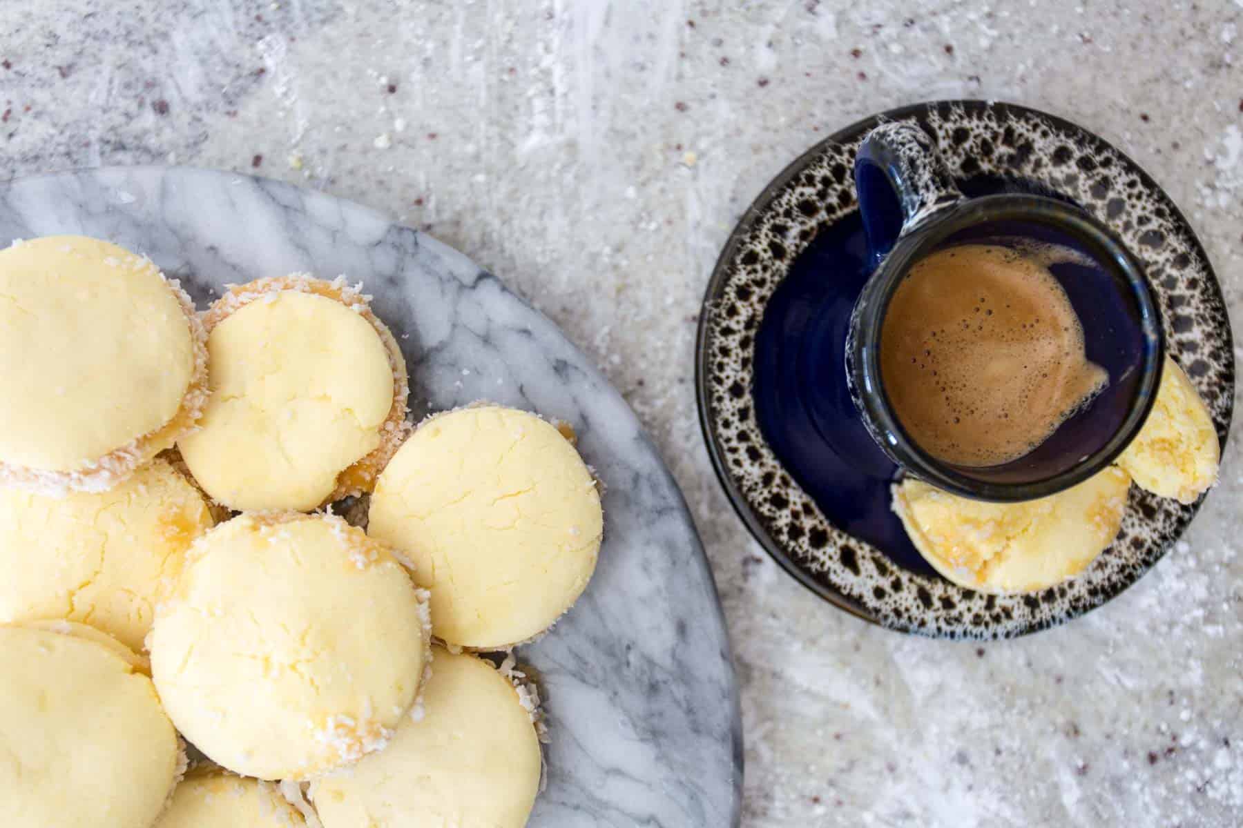 A marble platter with several round yellow cookies, reminiscent of gluten-free alfajores, is covered in shredded coconut beside a blue ceramic cup filled with coffee on a matching saucer. One cookie rests on the saucer's edge against the light speckled background.