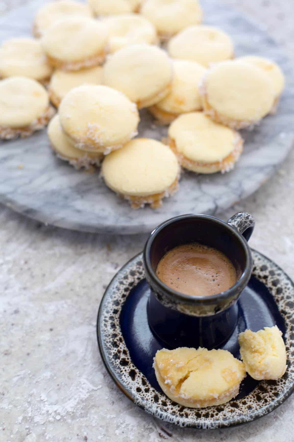 A dark blue espresso cup with a saucer holds coffee and a broken cookie. In the background, a marble tray showcases a pile of gluten-free alfajores with fillings and powdered edges, all set on a light speckled surface.