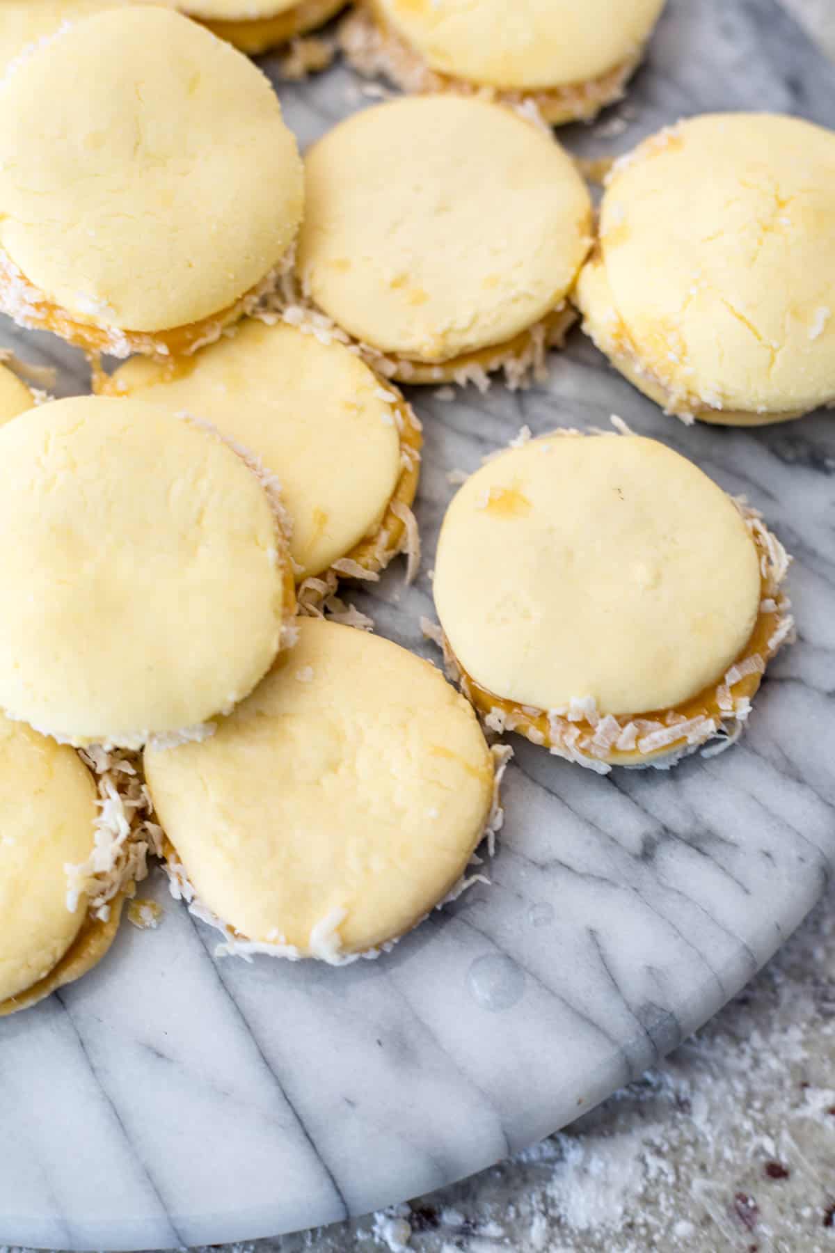 A pile of gluten-free alfajores, filled with caramel and rolled in shredded coconut, is displayed on a round, gray marble surface. The cookies boast a smooth, pale yellow outer layer.