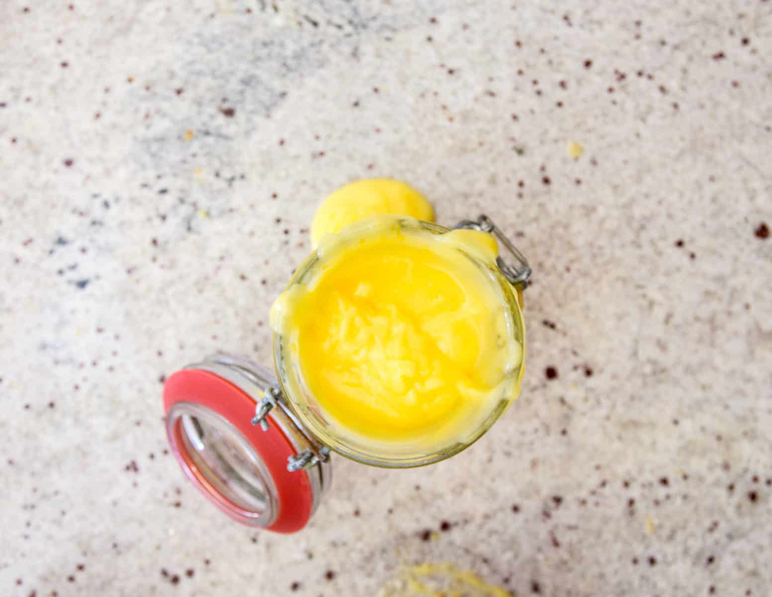 An open jar filled with creamy, yellow lemon curd rests on a speckled countertop beside gluten-free alfajores. The lid, attached with a metal hinge and featuring a red rim, lies slightly tilted to the side. Some lemon curd has spilled over the edge of the jar.