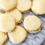 Close-up of a group of round, pale yellow gluten-free alfajores on a marble surface. The cookies have a filling and are partially coated with shredded coconut around the edges.