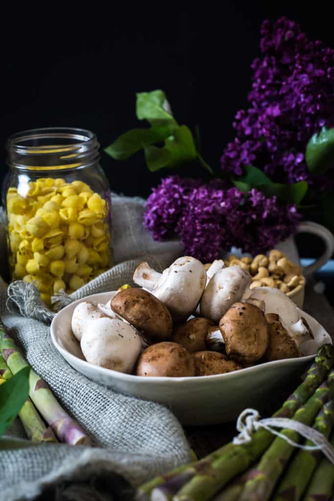 A vegan pasta primavera featuring mushrooms, asparagus, and purple flowers on a table.