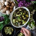 A hand serves a green vegetable and mushroom vegan pasta primavera from a white oval bowl into a smaller black bowl. Nearby are whole mushrooms in a white bowl, purple flowers, and green leafy branches on a wooden surface.