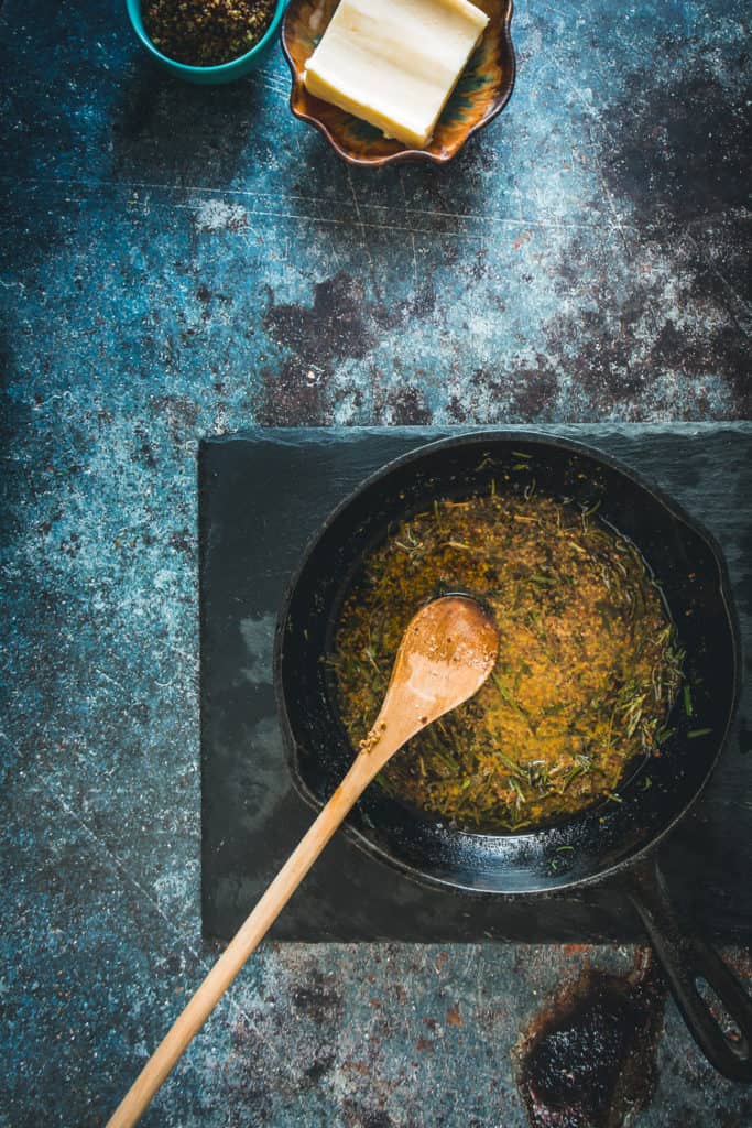 Herbs in melted butter cooking in cast iron pan with wooden spoon