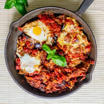 A black skillet contains eggplant shakshuka, a dish with poached eggs in a tomato and vegetable sauce, garnished with fresh basil leaves. The skillet is placed on a light-colored, woven mat background.