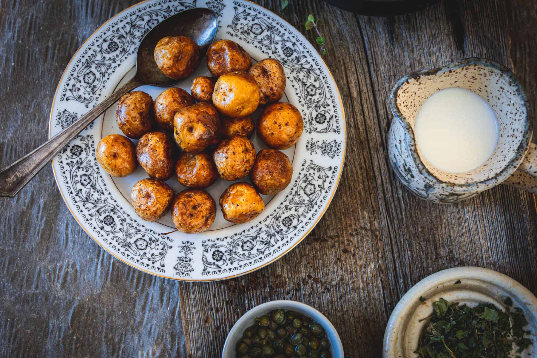 A plate of roasted potatoes sits on a wooden table, evoking the comforting vibe of an ajiaco colombiano. A spoon rests on the plate. Nearby, there's a ceramic jug with milk, a small bowl with capers, and another brimming with herbs, enhancing the rustic atmosphere.