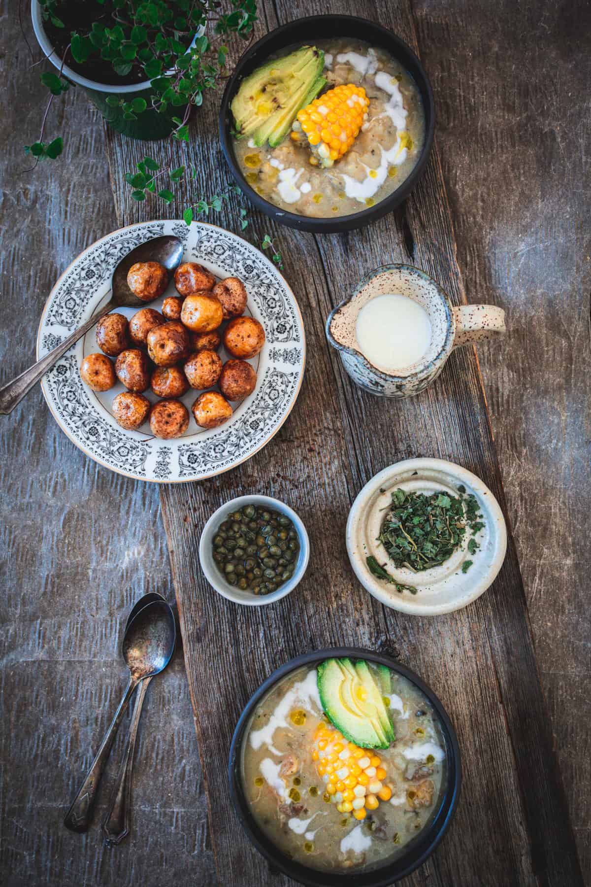 Two bowls of ajiaco colombiano soup with avocado, corn, and herbs sit on a wooden table. Nearby are seasoned potatoes, a bowl of capers, and dried herbs. A jug of milk and two metal spoons complete the scene, while a small plant pot peeks from the corner.