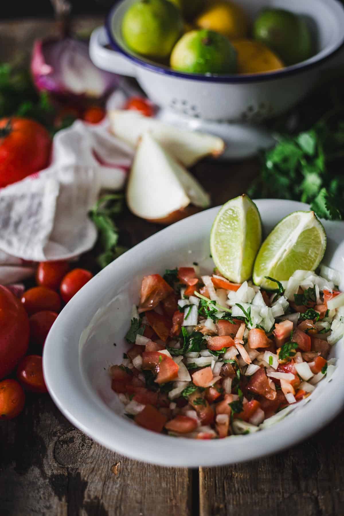 A bowl of freshly prepared Colombian empanada sauce sits in the foreground with diced tomatoes and herbs visible, accompanied by lime wedges; in the background, a rustic wooden table holds ingredients like whole tomatoes