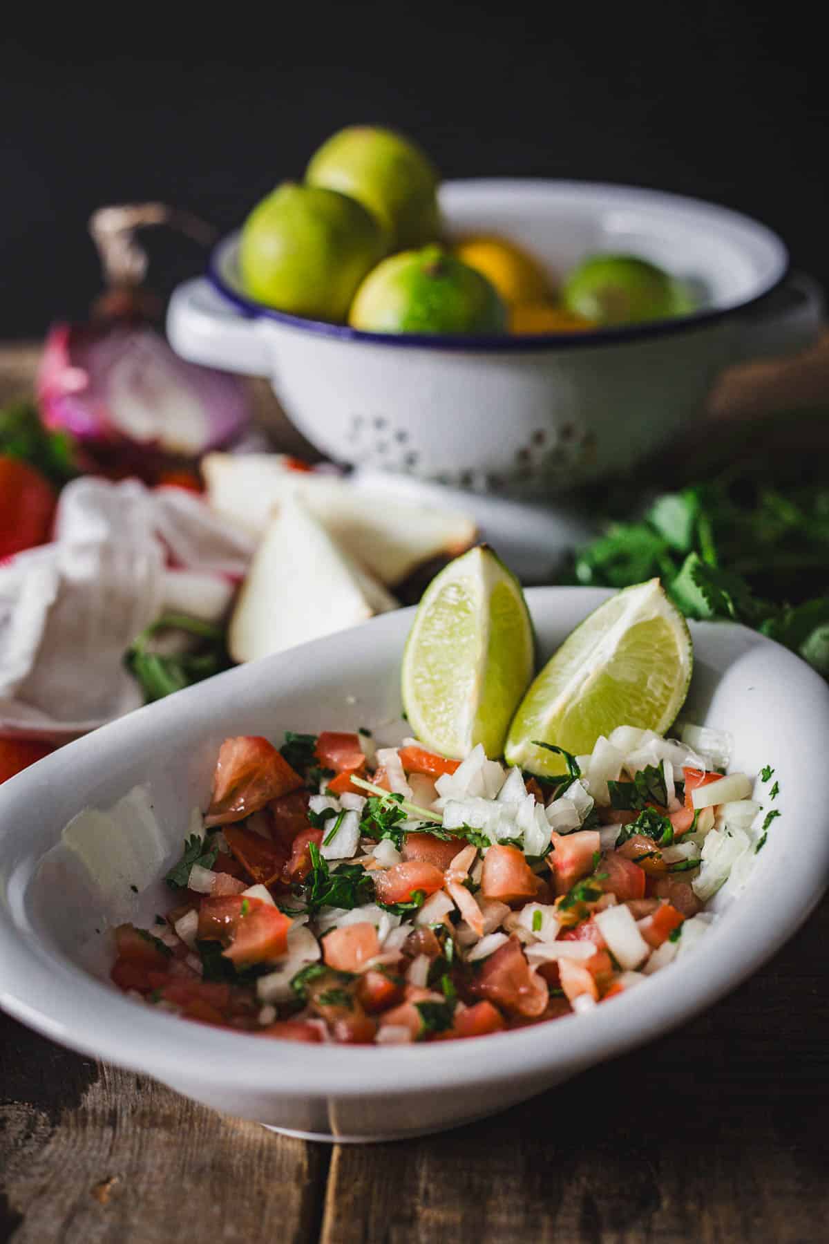 A plate of freshly made tomato salsa, intended as Colombian empanada sauce, garnished with chopped onions and cilantro, accompanied by lime wedges, with a colander of citrus fruits and vegetables