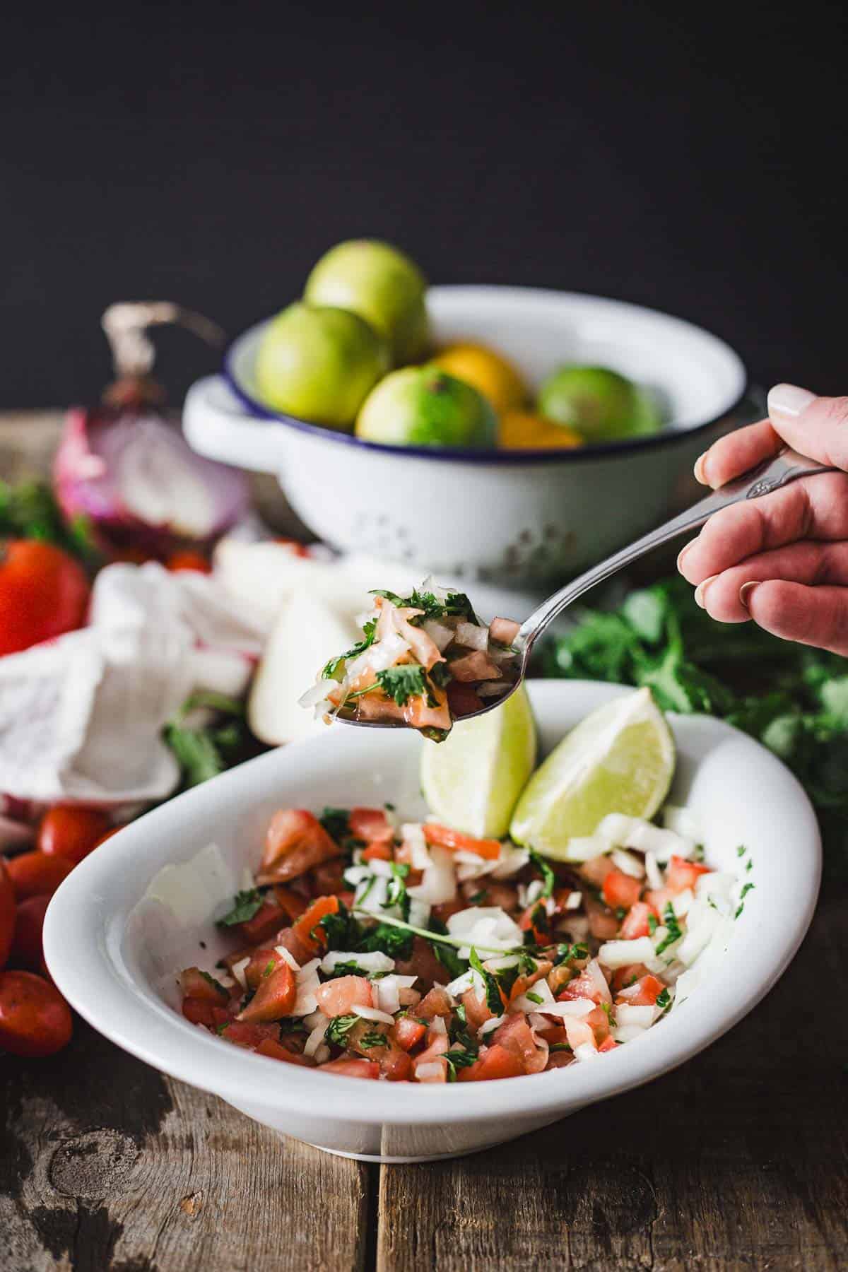 A hand holding a fork serves a portion of fresh Colombian empanada sauce with chopped tomatoes, onions, and herbs, garnished with lime wedges; ingredients are displayed in the background.