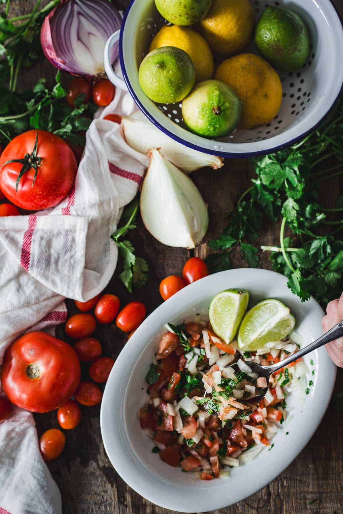 A bowl of fresh Colombian empanada sauce accompanied by a variety of whole vegetables and citrus fruits, suggesting the ingredients used in the preparation, presented on a rustic wooden surface.