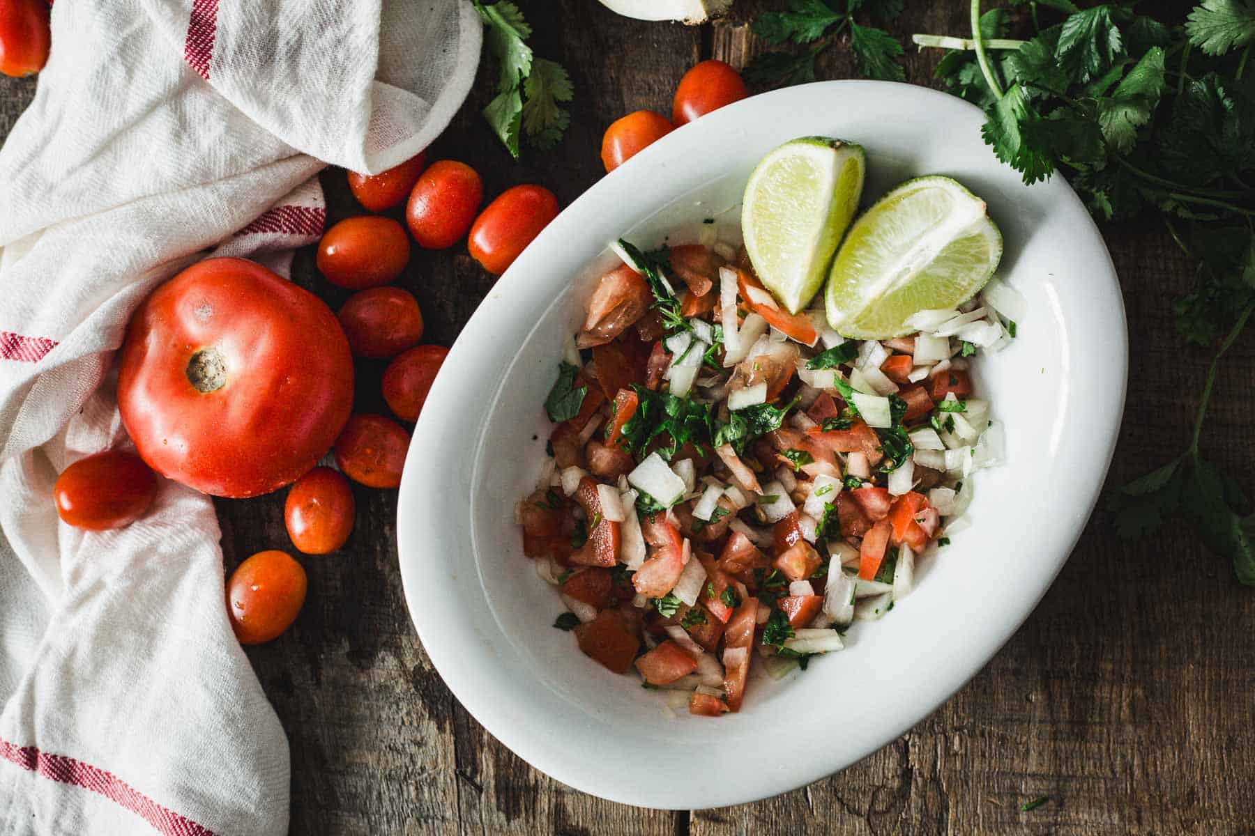 A bowl of freshly prepared Colombian empanada sauce garnished with lime wedges, surrounded by ingredients like whole tomatoes, cilantro, and cherry tomatoes on a wooden surface.