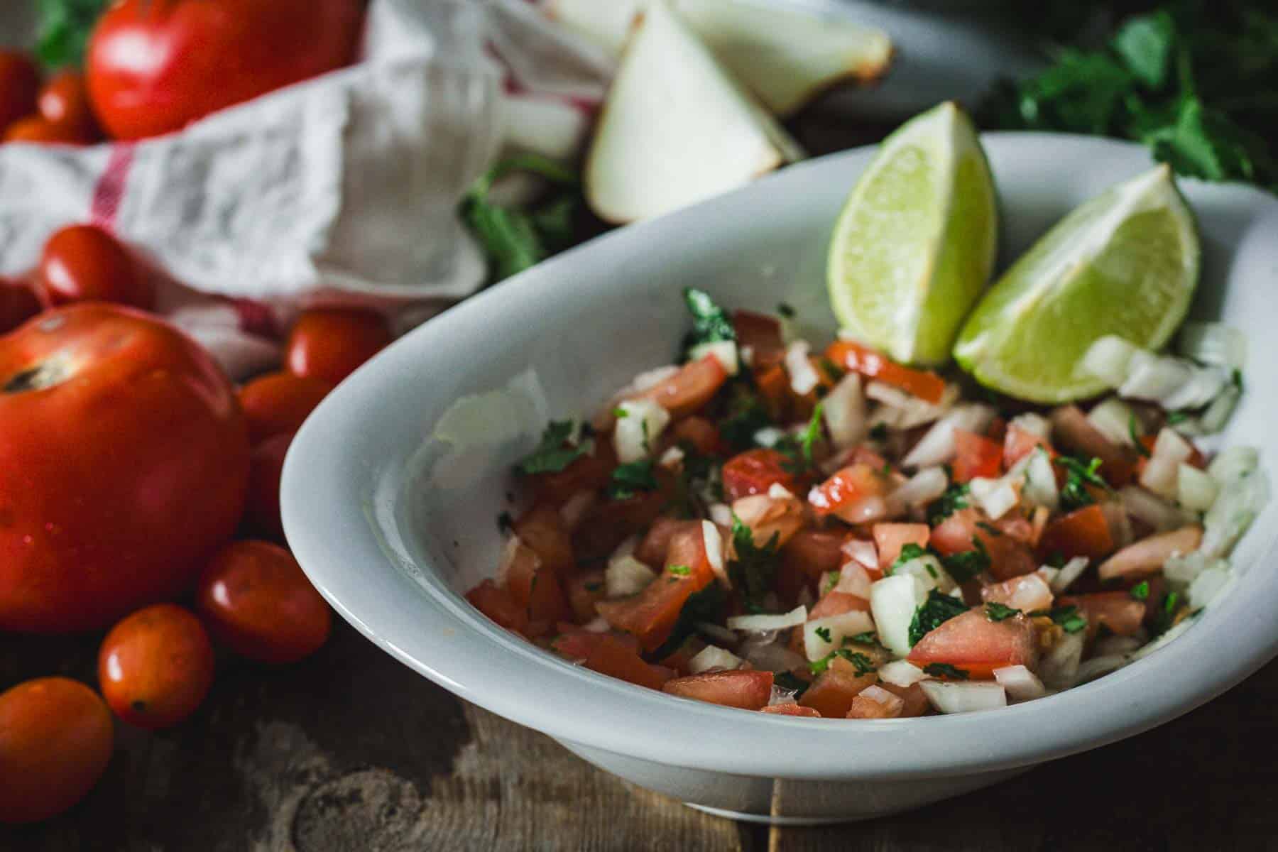 A bowl of Colombian empanada sauce with lime wedges on the side, surrounded by whole tomatoes, cherry tomatoes, onions, and fresh herbs on a wooden surface.