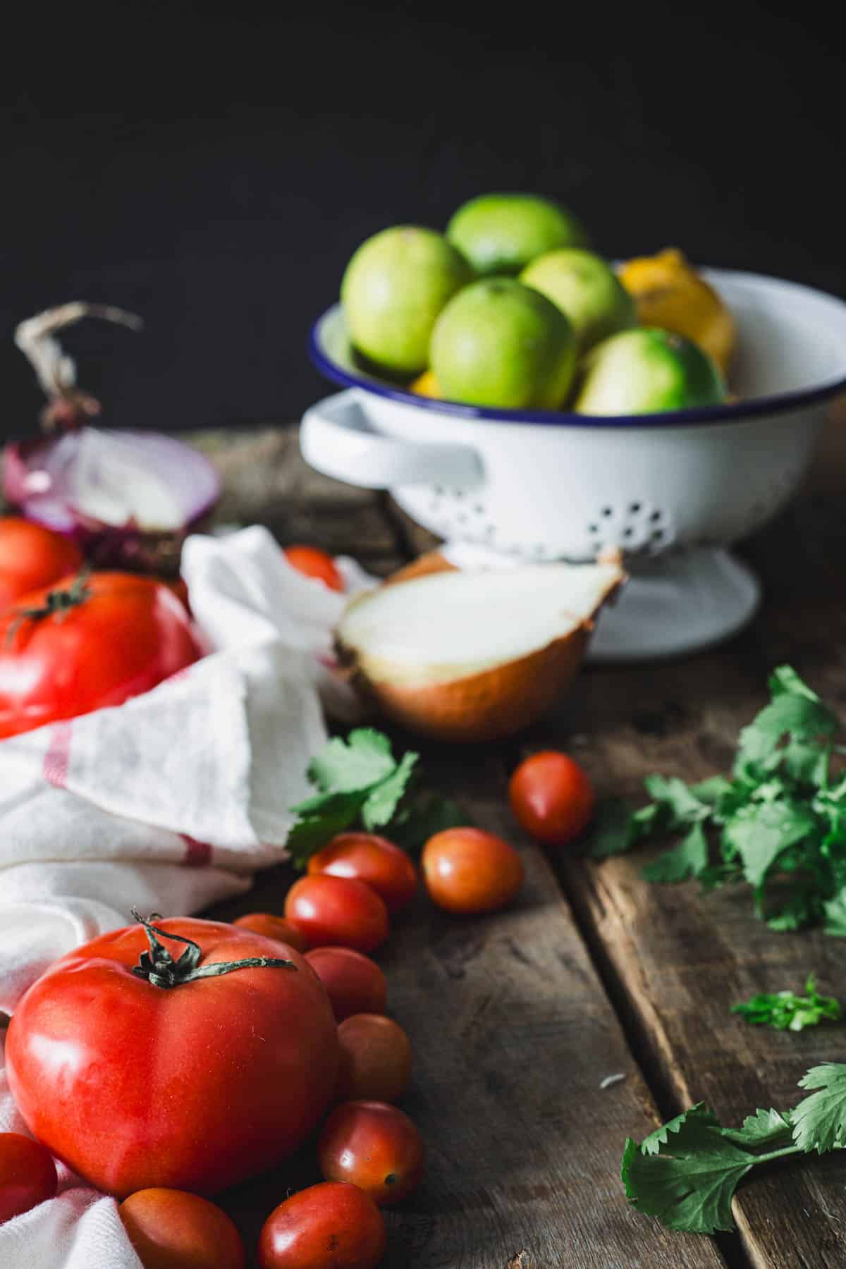 A variety of fresh produce, including tomatoes, green apples, and a cut onion, arranged on a wooden surface with Colombian empanada sauce and a white colander in the background.