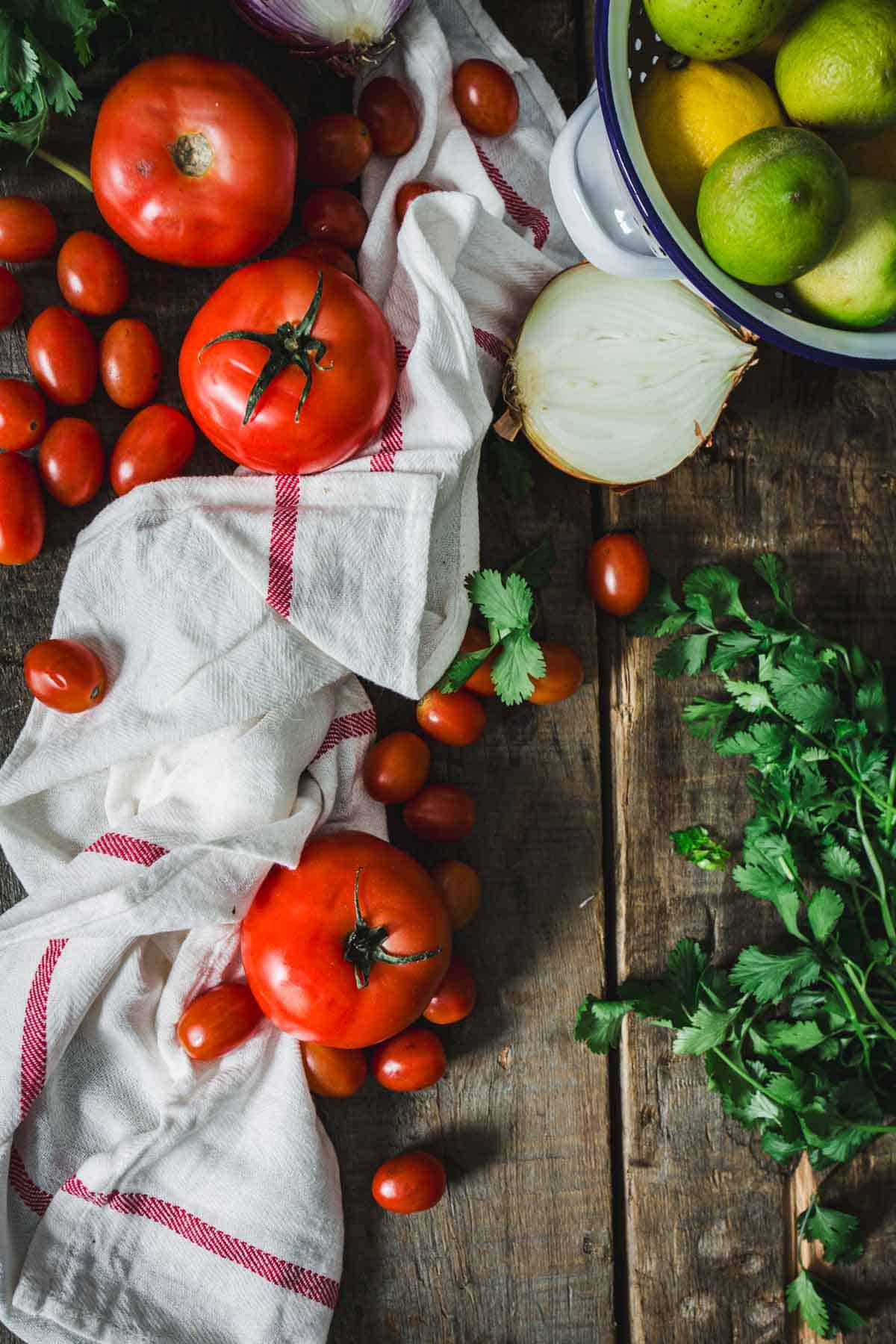 Various fresh vegetables and fruits, including tomatoes, onions, and citrus, arranged on a rustic wooden surface with a cloth napkin alongside Colombian empanada sauce.